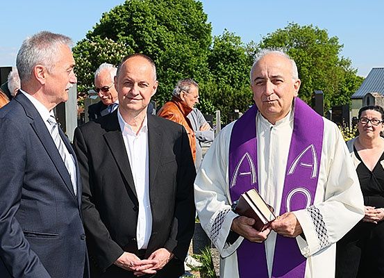 Ein Priester hält ein Buch, während zwei Männer in Anzügen neben ihm sprechen, mit einer Frau und einem älteren Mann im Hintergrund.
