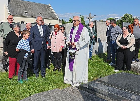 Ein Priester in einer weißen Robe hält einen silbernen Kelch auf einem Friedhof, wo Menschen in formeller Kleidung stehen, einige mit Brille.