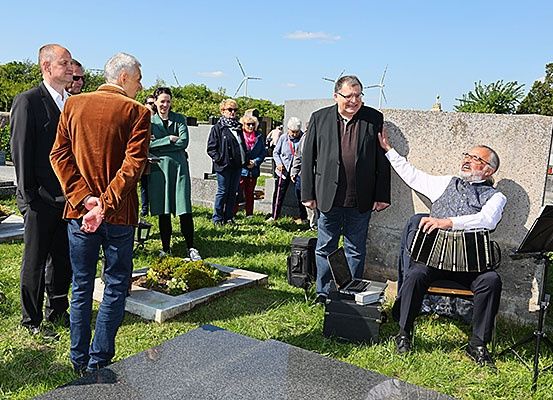 Eine Gruppe von Menschen steht auf einem Friedhof, wobei ein Mann auf einem Stuhl sitzt und Akkordeon spielt, während ein anderer Mann sich an einem Grabstein lehnt.