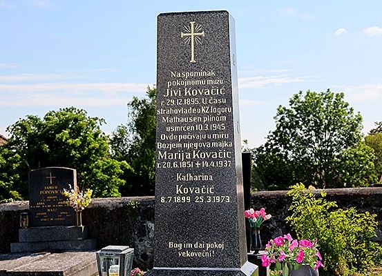 Ein Grabstein auf einem Friedhof mit einem Kreuz, Blumen und Inschriften in einer fremden Sprache. Ein weiterer Grabstein ist in der Nähe zu sehen, ebenfalls mit Blumen geschmückt.