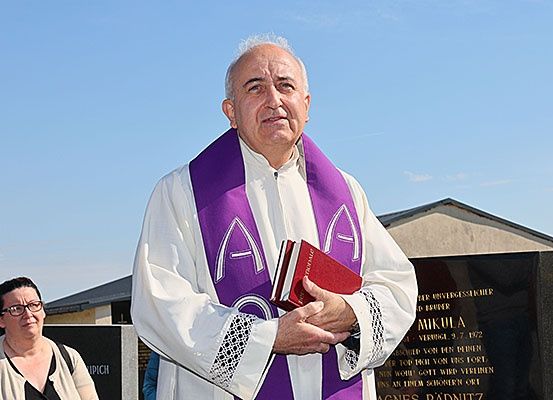 Ein Priester steht vor einem Denkmal, hält ein Buch in der Hand, mit einer Frau in der Nähe, die eine Brille trägt, unter einem klaren blauen Himmel.