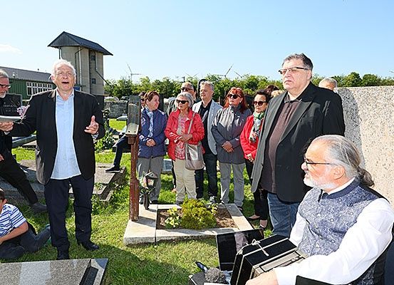 Eine Gruppe von Menschen auf einem Friedhof, ein älterer Mann spricht, ein anderer sitzt mit einem Akkordeon, ein kleiner Garten mit Pflanzen, eine Steinmauer und eine Windmühle in der Ferne.