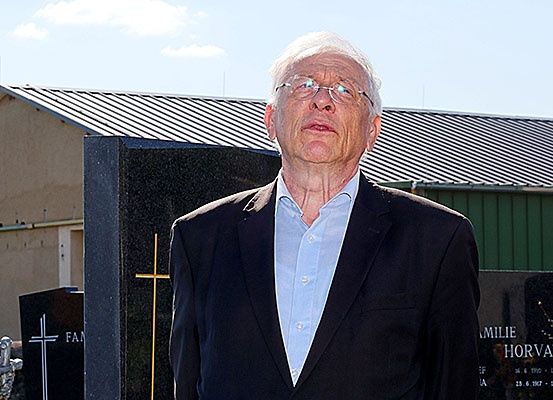 Ein älterer Mann in einem dunklen Anzug und Brille steht vor einem Grabstein mit einem Kreuz. Hinter ihm befindet sich ein Gebäude mit einem Metalldach.