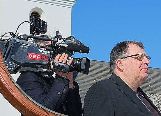 Ein Mann in einem Anzug, der eine große Kamera hält, filmt eine Person mit Brille, die vor einem Gebäude steht.