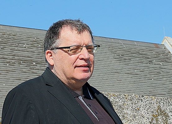 Ein Mann mit Brille steht vor einem Haus mit grauem Dach. Er trägt einen schwarzen Mantel und ein braunes Hemd. Er schaut nach rechts.