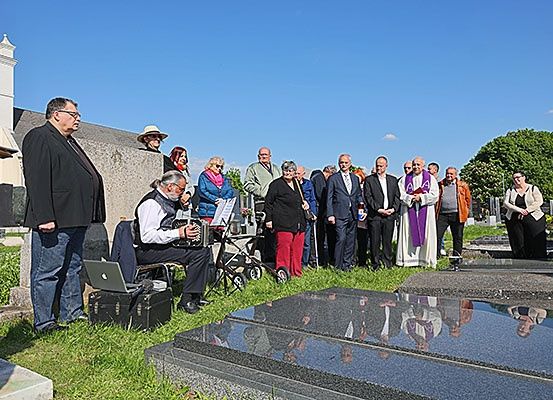 Eine Gruppe von Menschen versammelt sich auf einem Friedhof mit einem Priester und einem Mann, der Akkordeon spielt.