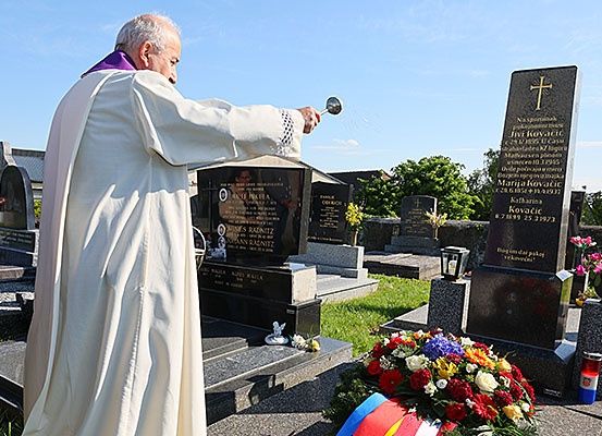 Ein Priester steht an einem Grab, gießt heiliges Wasser. Das Grab ist mit einem Kreuz und Blumen gekennzeichnet.