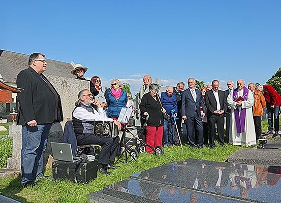 Eine Gruppe von Menschen versammelt sich auf einem Friedhof, wobei ein Priester vor ihnen steht. Ein Mann spielt Akkordeon, während er auf dem Gras sitzt.