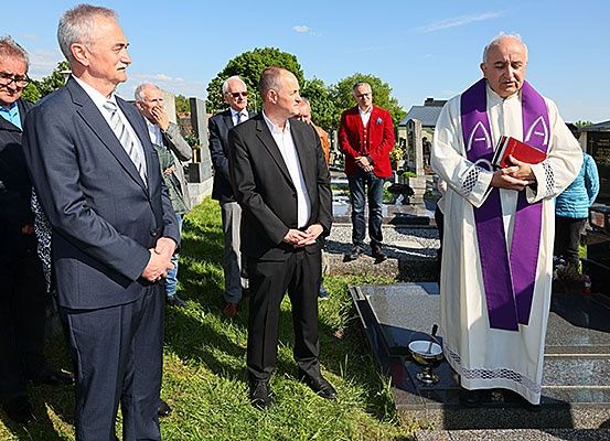 Ein Priester steht an einem Grab und liest aus einem Buch. Männer in Anzügen und ein Mann in einem roten Jackett stehen hinter ihm.