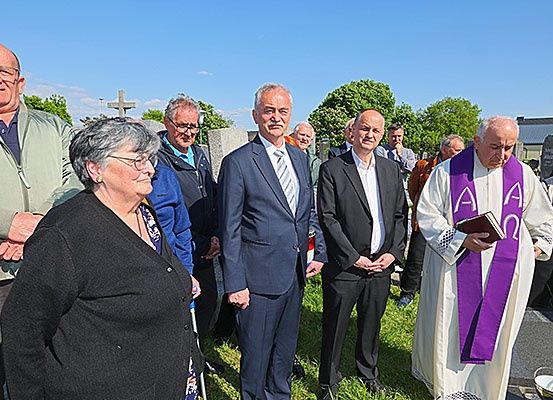 Eine Gruppe von Menschen, einschließlich eines Priesters, steht auf einem Friedhof, möglicherweise bei einer Beerdigung. Sie sind formell gekleidet.