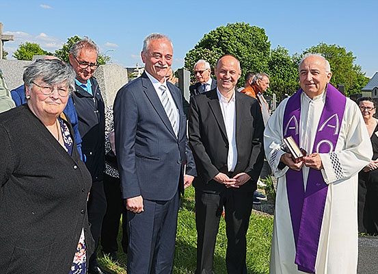 Eine Gruppe von Menschen, einschließlich eines Priesters, steht auf einem Friedhof. Einige sind formell gekleidet, andere in Freizeitkleidung. Bäume und ein Kreuz sind im Hintergrund.