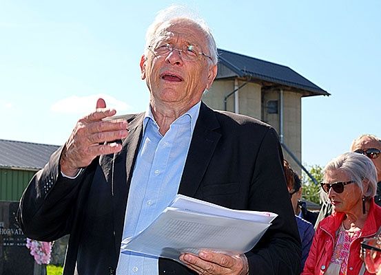 Ein älterer Mann mit Brille spricht und hält ein Papier in der Hand, möglicherweise auf einem Friedhof. Hinter ihm steht ein Grabstein mit dem Namen Horvat. Dahinter stehen weitere Menschen, und im Hintergrund befindet sich ein Gebäude.