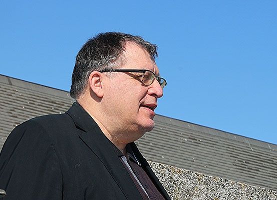 Ein Mann mit Brille steht im Freien in der Nähe einer Steinmauer und schaut unter einem klaren blauen Himmel nach oben.