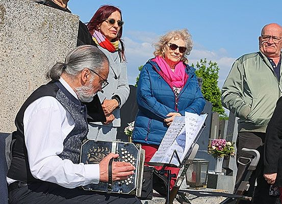 Ein älterer Mann spielt Akkordeon auf einem Friedhof, während mehrere Leute um ihn herumstehen.