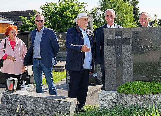Fünf Personen stehen auf einem Friedhof, wobei zwei zu den anderen sprechen. Ein Mann trägt einen Hut, und alle lächeln. Gräbersteine befinden sich hinter ihnen, wobei einer ein Kreuz und Text hat.