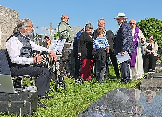 Eine Gruppe von Menschen versammelt sich auf einem Friedhof, wobei ein Mann ein Akkordeon spielt und ein anderes eine Musikpartitur in der Hand hält. Hinter ihm stehen mehrere Personen, darunter ein Priester in einer weißen Robe. Ein Laptop und eine schwarze Tasche liegen auf dem Boden.