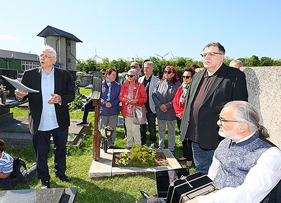 Eine Gruppe von Menschen versammelt sich auf einem Friedhof, wobei ein Mann die Gruppe anspricht und dabei ein Stück Papier in der Hand hält. Einige stehen, während andere auf dem Gras sitzen.