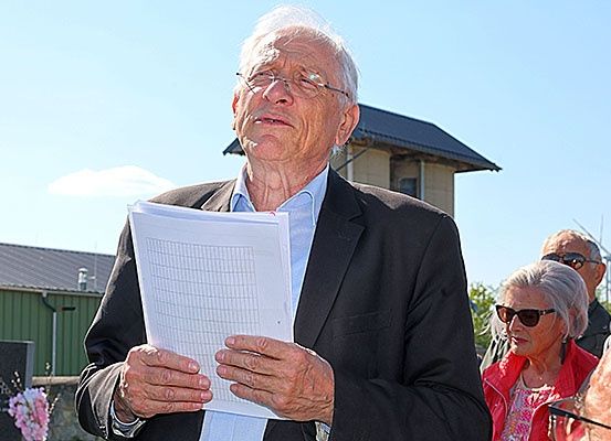 Ein älterer Mann hält ein Blatt Papier auf einem Friedhof in der Hand und schaut darauf. Hinter ihm steht eine Frau mit weißen Haaren, die eine Sonnenbrille trägt. Dahinter ist ein Gebäude mit einem schwarzen Dach zu sehen.