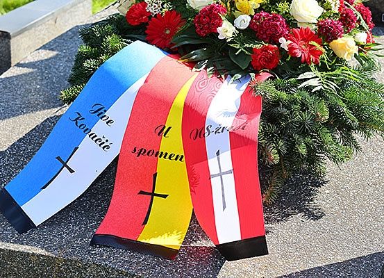 Ein Blumenkranz mit blauen, weißen, roten und gelben Bändern, jeweils mit einem Kreuz, ruht auf einem Steindenkmal.