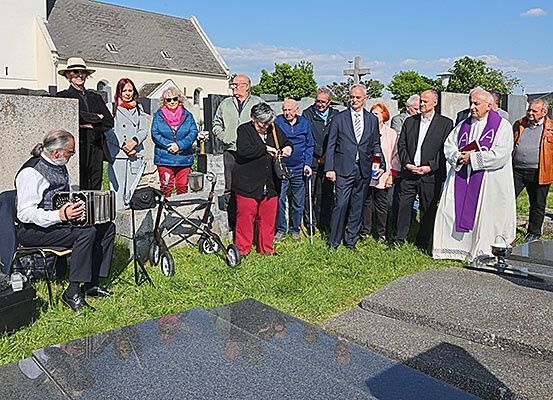 Eine Gruppe von Menschen versammelt sich auf einem Friedhof mit einem Priester vor einem Grab. Ein Mann spielt Akkordeon, während eine andere Frau einen Rollstuhl benutzt.