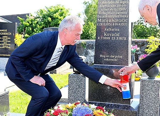 Zwei Männer in Anzügen platzieren eine Kerze auf einem Grab mit Blumen in der Nähe. Hinter ihnen ist ein Grabstein mit Text in einer fremden Sprache zu sehen.