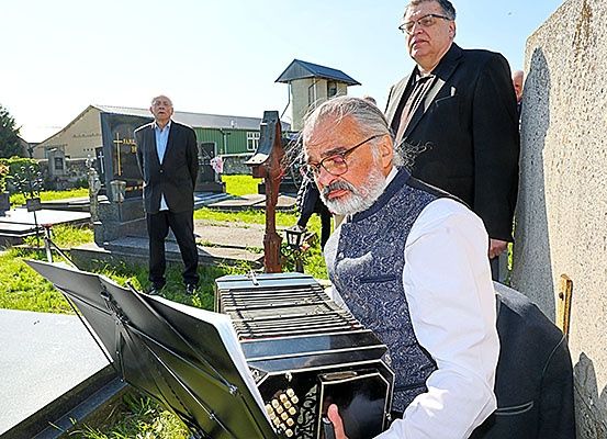 Ein älterer Mann spielt ein Akkordeon auf einem Friedhof, während zwei Männer in Anzügen im Hintergrund zusehen.