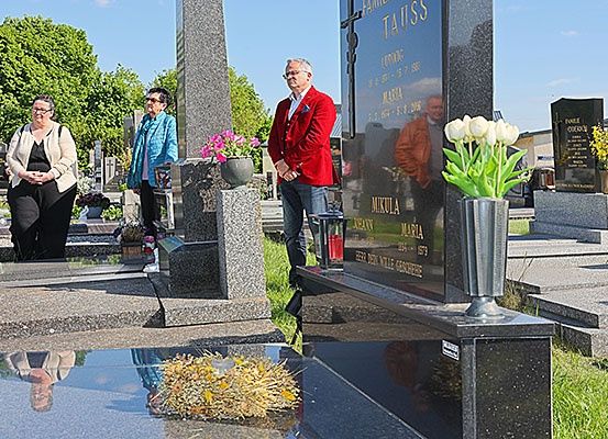 Eine Gruppe von Menschen steht auf einem Friedhof, ein Mann in einem roten Jackett, zwei Frauen in Jacken, eine mit Brille. Grabsteine mit Blumen und Inschriften sind um sie herum.