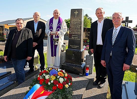 Fünf Männer stehen auf einem Friedhof, einer hält ein Buch, neben einem Grabstein mit einem Kreuz und Blumen. Sie sind formell gekleidet, und die Atmosphäre ist feierlich.