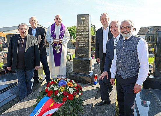 Sechs Männer in Anzügen stehen auf einem Friedhof, einer hält eine Trophäe. Ein Priester in einer Kutte steht in der Mitte und Blumen liegen auf dem Boden.