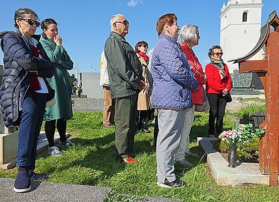 Eine Gruppe von Menschen steht auf einem Friedhof, vor einem Kreuz mit Blumen. Sie tragen Winterkleidung und wirken nachdenklich. Ein Kirchturm ist im Hintergrund zu sehen.
