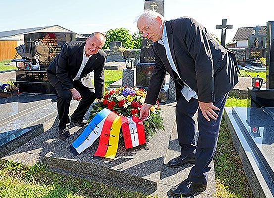 Zwei Männer in Anzügen platzieren eine Flagge und einen Kranz auf einem Grab in einem Friedhof.