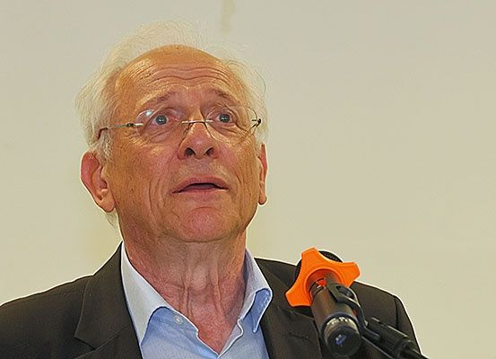 Ein älterer Mann mit weißen Haaren und Brille, gekleidet in einem schwarzen Anzug und blauem Hemd, spricht in ein Mikrofon.