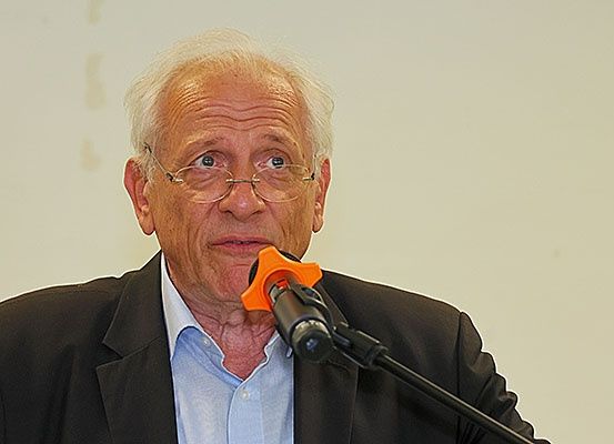 Ein älterer Mann mit einer Brille und einem schwarzen Jackett spricht in ein Mikrofon.