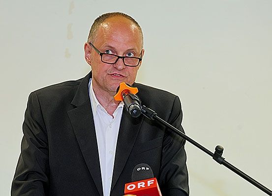 Ein Mann in einem schwarzen Anzug und mit Brille steht hinter einem Podium mit Mikrofonen. Er scheint eine Rede zu halten.