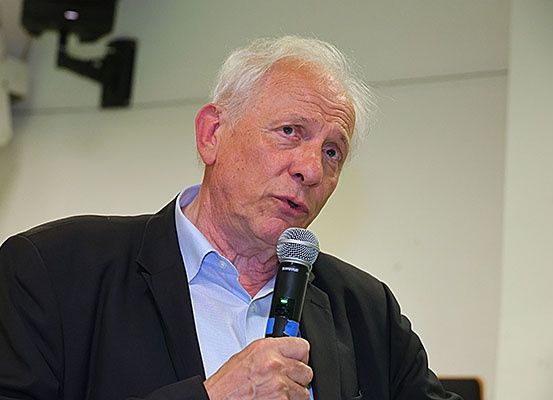 Ein älterer Mann spricht in ein Mikrofon, er trägt einen schwarzen Anzug und ein hellblaues Hemd.