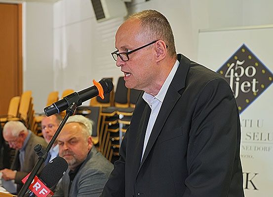 Ein Mann in einem schwarzen Anzug spricht in ein Mikrofon an einem Rednerpult. Hinter ihm sitzen mehrere Männer, einer mit Brille und ein anderer in einem Anzug. Im Hintergrund befindet sich ein Banner mit dem Text '450 jet'.