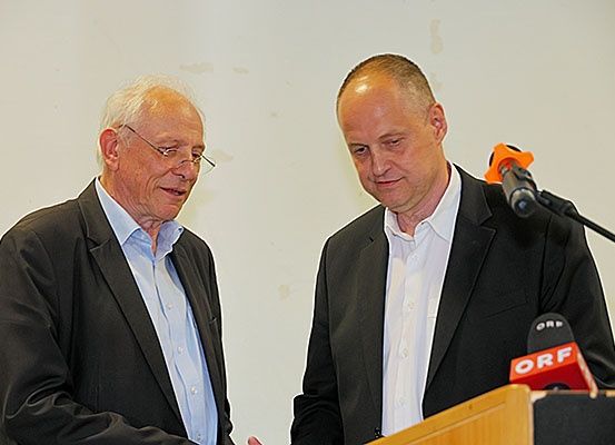 Zwei Männer in Anzügen stehen an einem Podium mit Mikrofonen, die mit ORF beschriftet sind. Ein Mann ist älter mit Brille, der andere ist jünger. Sie scheinen ein Gespräch zu führen.