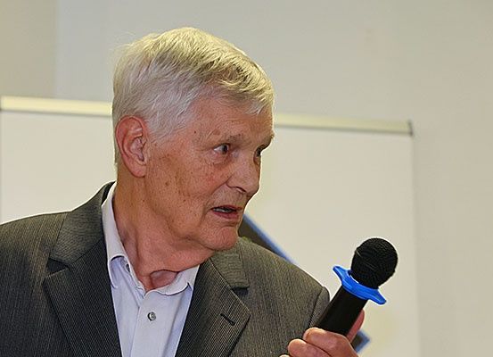Ein älterer Mann mit weißen Haaren hält ein Mikrofon. Er trägt einen grauen Anzug und ein weißes Hemd. Er scheint vor einer weißen Wand zu sprechen.