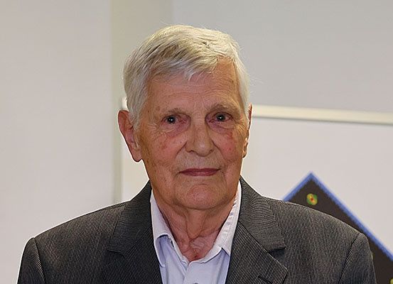 Ein älterer Mann mit weißem Haar und einem Lächeln posiert für ein Bild in einem Raum mit weißen Wänden und einem gerahmten Kunstwerk.