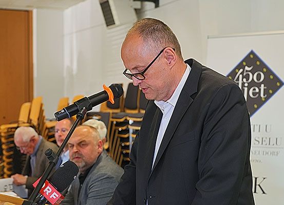 Ein Mann spricht in ein Mikrofon bei einer Konferenz. Hinter ihm steht ein Schild mit der Aufschrift '450 Jahre'. Andere Teilnehmer sitzen auf Stühlen.