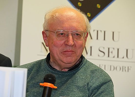 Ein älterer Mann mit Brille und einem grünen Pullover spricht in ein Mikrofon. Hinter ihm befindet sich eine Wand mit den Worten 'IMI SELU' und 'UDORF'.