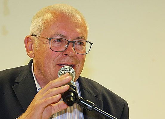 Ein älterer Mann mit Brille spricht in ein Mikrofon. Er hat ein leichtes Lächeln und ist in einem dunklen Anzug gekleidet.