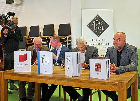 Fünf Männer sitzen an einem Tisch mit Büchern. Sie diskutieren wahrscheinlich etwas. Hinter ihnen filmt ein Kameramann. Ein Banner mit dem Text '450ljet' ist im Hintergrund.