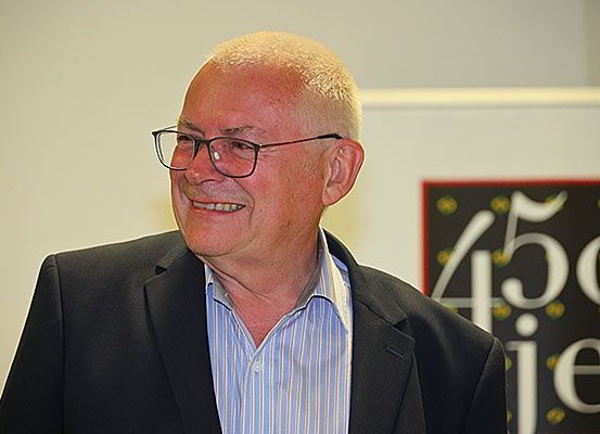 Ein Mann mit freundlichem Lächeln, der eine Brille und einen schwarzen Anzug trägt, steht vor einer weißen Wand. Hinter ihm befindet sich ein Schild mit dem Text 450 je.