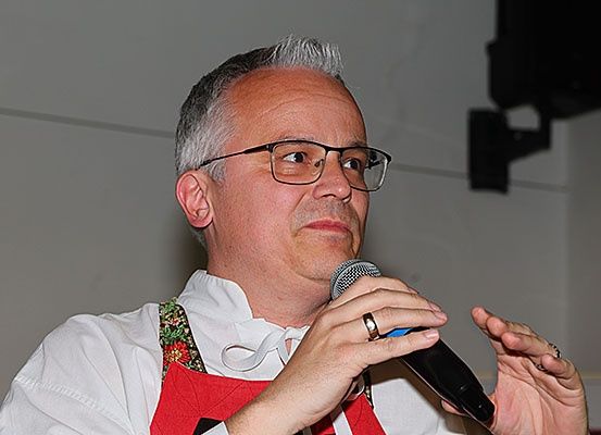Ein Mann in einem weißen Hemd und einer roten Schürze mit Blumenmuster hält ein Mikrofon in beiden Händen und scheint zu sprechen.