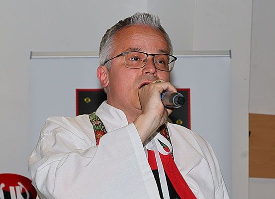 Ein Mann mit Brille und einem weißen Hemd mit rotem Band spricht in ein Mikrofon. Die Wand hinter ihm hat ein rotes und schwarzes Design.