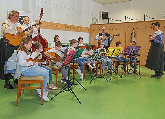 Eine Gruppe von Kindern in einem Raum spielt Gitarren und Celli mit zwei Erwachsenen, die hinten stehen.