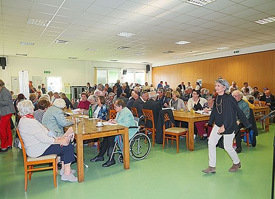 Ein Raum mit Tischen und Stühlen, in dem Menschen sitzen und reden. Eine Frau in weißen Hosen geht vor ihnen.