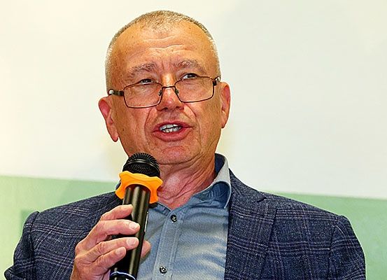 Ein älterer Mann mit Brille, der ein blaues Hemd und einen Blazer trägt, hält ein Mikrofon und scheint zu sprechen.