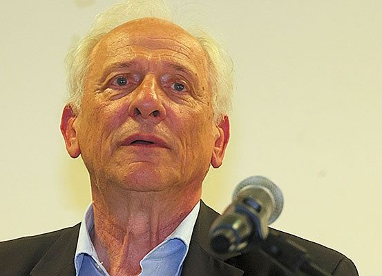 Ein älterer Mann mit weißen Haaren spricht in ein Mikrofon in einem Konferenzraum. Er trägt ein blaues Hemd und einen schwarzen Anzug.